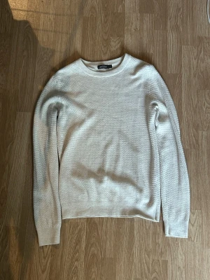 Vit stickad tröja med strukturmönster - Säljer en stilren vit stickad tröja med diskret strukturmönster och rund hals. Tröjan har långa ärmar och ribbade muddar vid ärmslut och nederkant. Perfekt för lager på lager och enkel att matcha med olika outfits.