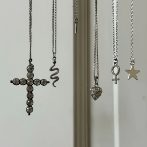 Silver halsband - Sex halsband i silverfärg med olika hängen: ett kors med pärlor, en orm, bokstaven I, ett hjärta med stenar, en venus-symbol och en stjärna. Kedjorna är tunna och smyckena har en modern och stilren känsla.
