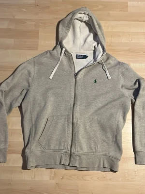 Grå hoodie från Polo Ralph Lauren - Klassisk grå hoodie från Polo Ralph Lauren med dragkedja. Perfekt för en avslappnad och stilren look.