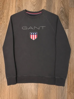 Mörkgrå sweatshirt från GANT - Snygg mörkgrå sweatshirt från GANT med broderad logga och sköld med stjärnor och ränder på bröstet. Tröjan har rund halsringning och ribbade muddar vid ärmslut och nederkant. Perfekt för en avslappnad och stilren look.