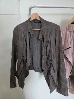 Grå skinnjacka från Jus d'Orange  - Snygg grå skinnjacka från Jus d'Orange med asymmetrisk nederkant och dekorativa dragkedjor framtill. Jackan har hög krage med knappdetaljer och en cool, avslappnad passform. Perfekt för dig som vill ha en edgy look.