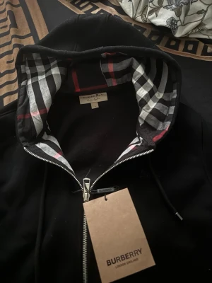  Burberry hoodie - Snygg svart hoodie från Burberry med dragkedja och klassiskt rutigt foder i huvan. Hoodien har dragsko i huvan och diskret Burberry-logga broderad på bröstet. Perfekt för dig som vill ha en stilren och lyxig look.