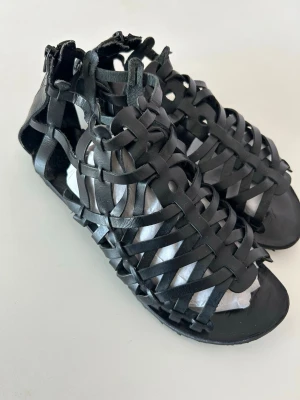 Svarta flätade lädersandaler - Snygga svarta sandaler i flätad design med öppen tå och häl. Tillverkade i läder med dragkedja bak för enkel påtagning. Perfekta för en trendig och luftig look under varma dagar.