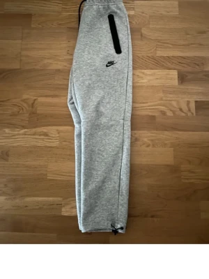 Grå mjukisbyxor från Nike - Snygga grå mjukisbyxor från Nike med svarta dragkedjor på fickorna och justerbar dragsko i midjan. Byxorna har även dragsko vid bensluten och Nike-logga broderad på låret. Perfekta för en avslappnad stil.