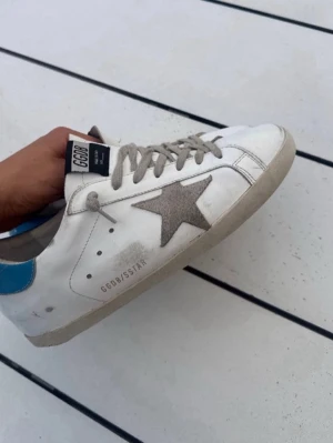 Golden Goose Skor med blå detaljer  - Golden Goose sneakers i vitt läder med grå stjärndetalj på sidan, grå skosnören och blå hälflik. Klassisk låg modell med GGDB/SSTAR-tryck på sidan och slitna detaljer för en cool, avslappnad look.
