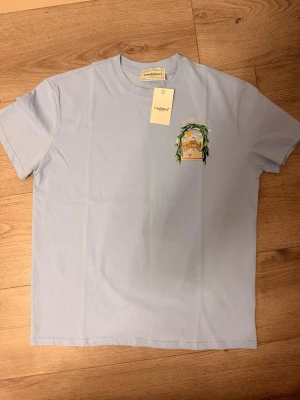 Ljusblå t-shirt från Casablanca - Ljusblå t-shirt från Casablanca med tryckt motiv på bröstet som föreställer en byggnad och gröna blad. Klassisk passform med rund hals och korta ärmar. Tillverkad i mjuk bomull för en skön känsla.