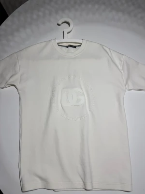 Vit t-shirt från Dolce & Gabbana - Vit t-shirt från Dolce & Gabbana med DG-logga och text i relief på bröstet. Klassisk rund halsringning och korta ärmar. Snygg och stilren design som passar till det mesta.