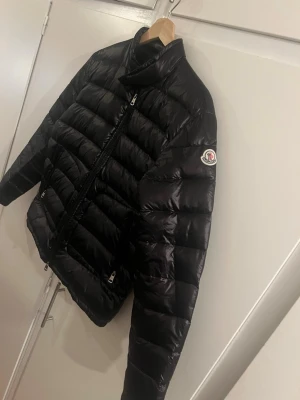 Svart dunjacka från Moncler - Snygg svart dunjacka från Moncler med klassisk quiltad design och hög krage. Jackan har dragkedja framtill, två fickor med dragkedja och Moncler-logga på ärmen. Perfekt för kalla dagar och riktigt stilren look. Kan även passa S användare fast den är storlek 2 
