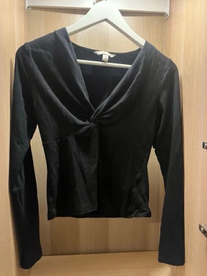 Svart twistad topp från H&M. - Snygg svart långärmad topp från H&M med twistad detalj framtill och djup v-ringning. Figurnära passform och mjukt material som känns skönt mot huden.