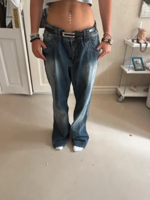 Baggy blå jeans med låg midja - Säljer ett par baggy blå jeans med låg midja och bred passform. Jeansen har slitna detaljer och ljusare partier framtill för en cool vintage-look. Klassisk femficksmodell och tillverkade i denim. Perfekta för en avslappnad streetstil.