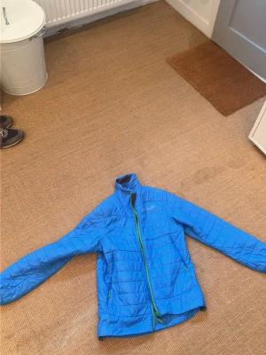 Blå pufferjacka från Norrøna - Snygg blå pufferjacka från Norrøna med grön dragkedja och hög krage. Jackan är lätt vadderad och har två sidofickor med dragkedja. Perfekt för kalla vinterdagar och har en sportig look som passar till det mesta.otroligt fint skick