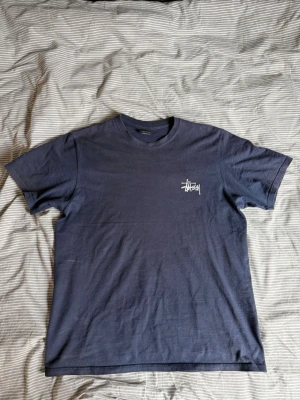 Mörkblå Stüssy t-shirt med logga - Säljer en mörkblå t-shirt från Stüssy med vit logga tryckt på bröstet och stor logga på ryggen. Klassisk passform och rund halsringning. Perfekt för dig som gillar streetwear och vill ha något stilrent men ändå med attityd.