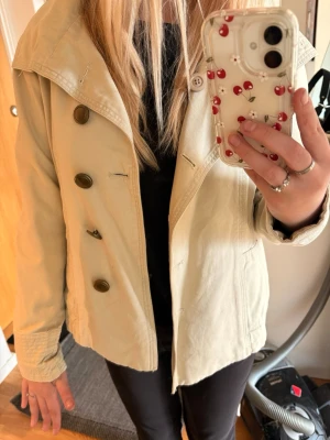 Trenchcoat  - Kort Beige trenchcoat men ett band rund midjan man kan justera, både snyggt utan som med band. Storlek S