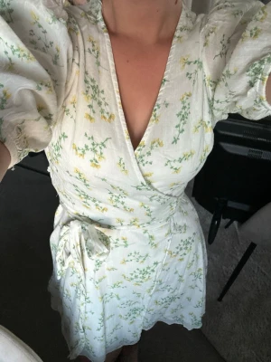 Vit omlottklänning med blommor - Söt vit omlottklänning med blommor. Färgen är som på första bilden men passformen syns bättre på andra! Endast använd vid ett par enstaka tillfällen så finns inga tecken på användning. Är storlek m men passar även s skulle jag säga!