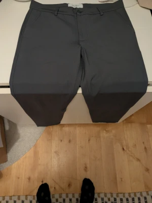 Mörkgrå kostymbyxor från Les Deux - Snygga mörkgrå kostymbyxor från Les Deux med klassisk passform och diskreta fickor. Materialet är lätt och bekvämt, perfekt för dig som vill ha en stilren look. Byxorna har hällor för bälte och stängs med knapp och dragkedja.