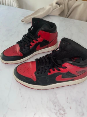 Air Jordan 1 Mid Bred Svart/Röd - Snygga Air Jordan 1 Mid sneakers i klassisk svart och röd färg med vit sula. Skorna har ikonisk Nike Swoosh och Wings-logga på sidan. Tillverkade i läder med svarta skosnören och perforerad tå för extra ventilation. Perfekta för dig som gillar streetwear och basketstil.