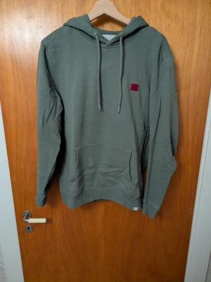Olivgrön hoodie från Les Deux - Snygg olivgrön hoodie från Les Deux med huva och dragsnören. Har en röd broderad patch med romerska siffran II på bröstet och en stor magficka framtill. Perfekt för en avslappnad och trendig stil.