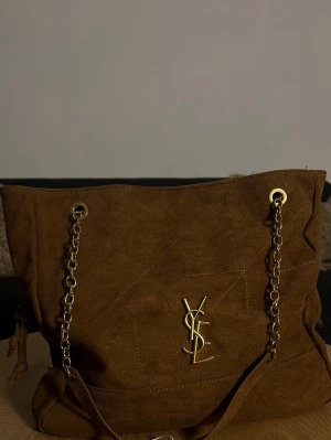 Brun handväska från YSL - Snygg brun handväska  från YSL med guldiga kedjehandtag och ikonisk YSL-logga framtill. Väskan har en mjuk, slouchy form och detaljer i guldmetall. Perfekt för dig som vill ha en trendig och lyxig accessoar.