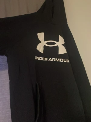Svart hoodie från Under Armour - Svart hoodie från Under Armour med stor vit logga och text på bröstet. Klassisk modell med huva och känguruficka framtill. Perfekt för en sportig och avslappnad stil. Mjuk och bekväm bomullsblandning.