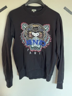 Svart Kenzo sweatshirt med tigertryck - Cool svart sweatshirt från Kenzo med ikoniskt broderat tigerhuvud och färgglada detaljer framtill. Tröjan har rund hals, långa ärmar och ribbade muddar. Perfekt statement-plagg för dig som gillar streetstyle och unika prints.