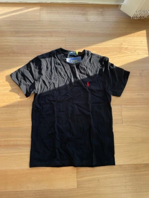 Svart Ralph Lauren T-shirt  - 👕 Märke: Polo Ralph Lauren 📏 Storlek: L 🎨 Färg: Svart med röd logga ✨ ☀️ Stilren och snygg t-shirt från Polo Ralph Lauren – perfekt för sommaren och enkel att matcha med både shorts och jeans. Tillverkad i mjuk och bekväm bomull med klassisk passform och ikonisk röd logga på bröstet.  ✨ Skick: Helt ny med tags kvar 🧼 Aldrig använd 👌 Tidlös modell som passar till allt