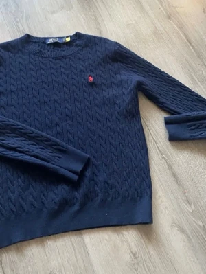 Mörkblå kabelstickad tröja Polo Ralph Lauren - Kabelstickad mörkblå tröja från Polo Ralph Lauren med rund halsringning och röd broderad logga på bröstet. Tröjan har långa ärmar och ribbade muddar vid ärmslut och nederkant. Perfekt för lager-på-lager och klassisk stil.