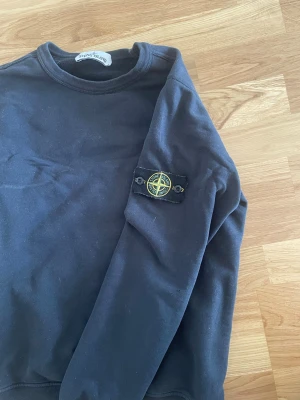 Svart sweatshirt från Stone Island - Cool svart sweatshirt från Stone Island med klassiskt rund hals och ribbade muddar. Ikonisk logopatch på vänster ärm som ger en snygg detalj. Perfekt för dig som gillar streetwear och vill ha något stilrent. Pris ej hugget i sten