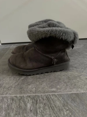 Gråa bailey bow mini UGGS - Mysiga gråa UGG boots med mjukt fluff intill. Klassisk modell med rund tå och grov sula för extra komfort. Perfekta för kalla dagar och ger en avslappnad look. UGG-logga bak på hälen. Liten fraktur framtill men inget man tänker på❣️pris går att diskutera 