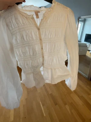 Vit blus med volanger från Nelly - Söt vit blus med volangkant nedtill och smockdetaljer över hela plagget. Blusen har knäppning framtill och långa ärmar med lätt puff. Tillverkad i ett tunt, luftigt tyg som ger en romantisk känsla. Perfekt att styla med jeans eller kjol.