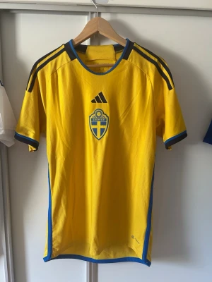 Sverige fotbollströja Adidas gul/blå - Gul fotbollströja från Adidas med Sveriges landslagsemblem på bröstet och svarta Adidas-ränder på axlarna. Blå detaljer vid ärmslut och sidor, samt texten 'Sverige' i nacken. Perfekt för match eller träning.