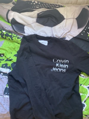 Svart sweatshirt från Calvin Klein Jeans - Svart sweatshirt från Calvin Klein Jeans med rund halsringning och tryckt logga i vitt och grått på bröstet. Klassisk modell med ribbade muddar vid hals, ärmslut och nederkant. Perfekt för en avslappnad och stilren look.