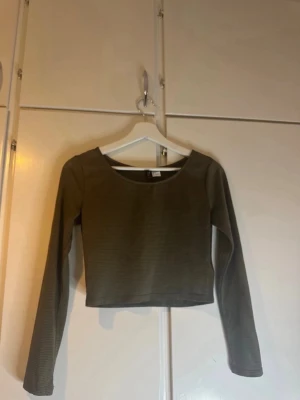 Olivgrön ribbad croppad topp Divided - Snygg olivgrön croppad topp från Divided med diskreta ränder och lång ärm. Toppen är ribbad och har en rund halsringning, perfekt att styla med jeans eller kjol för en trendig look.