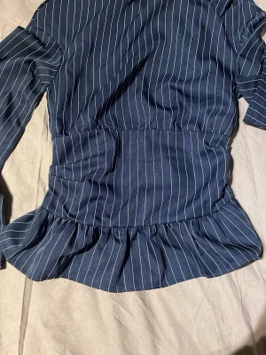 Blå randig blus med volang - Snygg blå blus med vita vertikala ränder och volang nertill. Modellen har lång ärm och markerad midja som ger en cool siluett. Perfekt att styla med jeans eller kjol för en trendig look.