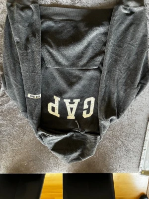 Svart hoodie från GAP med tryck - Mysig svart hoodie från GAP med stort vitt GAP-tryck på bröstet och en klassisk känguruficka framtill. Huvan har dragsko och är perfekt för en avslappnad stil. Tillverkad i mjukt material som känns skönt mot huden. Snygg detalj med 'EST. 1969' på ärmen.