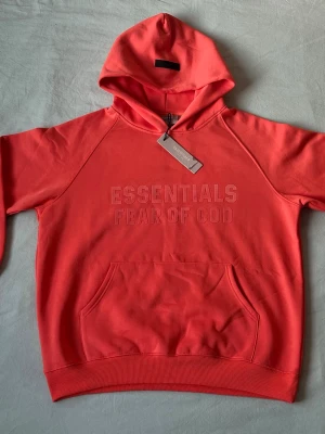 Koralröd Essentials Fear of God hoodie - Snygg korallröd hoodie från Fear of God Essentials med broderad logga på bröstet. Klassisk känguruficka framtill, huva och ribbade muddar. Perfekt för dig som gillar streetwear och vill sticka ut med en färgstark look.