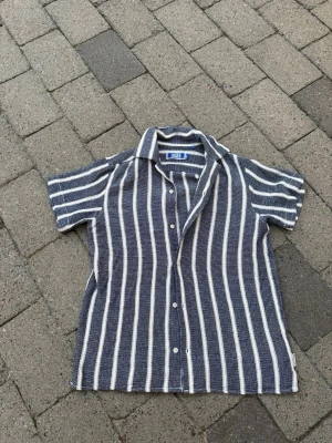 Randig skjorta från Jack & Jones - Snygg kortärmad skjorta från Jack & Jones med blå och vita vertikala ränder. Skjortan har klassisk krage, knäppning framtill och är tillverkad i ett strukturerat bomullstyg. Perfekt för en avslappnad och trendig look.
