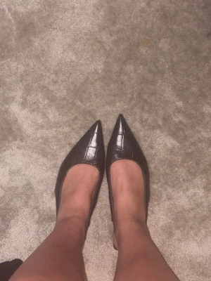 Bruna spetsiga ballerinaskor i skinn - Snygga bruna ballerinaskor med spetsig tå och croco-mönstrad yta. Skorna har öppen häl med rem och är tillverkade i ett skinnliknande material. Perfekta för dig som gillar trendiga och stilrena skor med en twist.