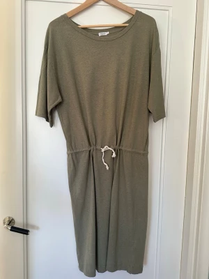 Olivgrön klänning från Filippa K - Olivgrön t-shirt klänning från Filippa K med avslappnad passform, rund halsringning och knytband i midjan. Klänningen har korta ärmar och är tillverkad i mjuk bomull, perfekt för en chill och stilren look. Observera ett litet hål vid skärpet samt sömmar dom släppt ngt men som inte påverkar fållen. 