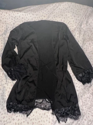 Svart morgonrock med spetsdetaljer - Snygg svart morgonrock i mjukt material med eleganta spetsdetaljer vid ärmslut och nederkant. Tillverkad av 95% polyester och 5% elastan för en bekväm och följsam känsla. Perfekt för mysiga hemmakvällar. 