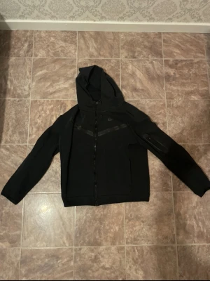 Svart Nike hoodie med dragkedja - Snygg svart hoodie från Nike med hel dragkedja framtill och huva. Diskreta svarta detaljer över bröstet och en ficka med dragkedja på ena ärmen. Perfekt för en sportig och avslappnad stil.