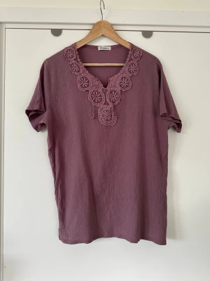 Lila blus med spetsdetaljer från Elisai, storlek L - Luftig fin rosa / lila blus / topp från Elisai med kort ärm och V-ringning med lätt boho-inspirerad design. Blusen har vackra broderade (virkade?) detaljer runt halsen och en avslappnad passform. Nedtill finns små sprund i sidorna. (Det står storlek 4XL men är en vanlig L). Mycket fint skick, som ny.