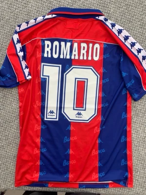 Barcelona Kappa Romario tröja - Retro Barcelona matchtröja från Kappa med Romario och nummer 10 på ryggen. Tröjan har klassiska blå och röda vertikala ränder, klubbmärke på bröstet och Kappa-loggor på ärmarna. Krage i blått och rött för extra stilpoäng. Perfekt för dig som älskar vintage fotbollströjor.