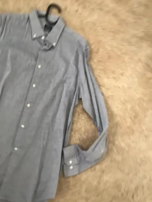 Blå långärmad skjorta med knappar - Klassisk blå skjorta med lång ärm och vita knappar. Skjortan har button-down krage och är tillverkad i ett lätt bomullstyg. Perfekt för en stilren och enkel look.