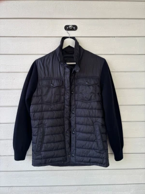 Massimo Dutti Cardigan - Snygg svart quiltad pufferjacka med långärmade stickade ärmar och hög krage. Jackan har två bröstfickor med knappar och två sidofickor med dragkedja. Perfekt för kyliga dagar och enkel att matcha med olika outfits.