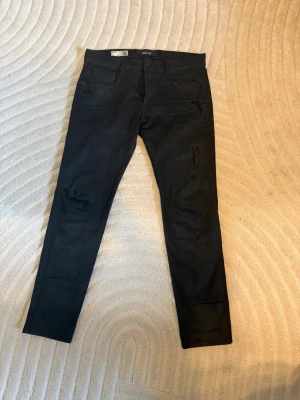 Svarta slim fit jeans från Replay - Säljer ett par svarta slim fit jeans från Replay, modell Anbass. Jeansen har klassisk femficksdesign, diskreta slitningar på benen och dragkedjegylf. Perfekta för en stilren och modern look.