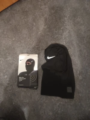 Nike Pro Therma-FIT svart huva - Nike Pro Therma-FIT Hood i svart, tillverkad i ett mjukt och stretchigt material som håller värmen. Huvan har en vit Nike-logga framtill och är designad för att sitta tight runt huvudet. Perfekt för träning utomhus eller kalla dagar.