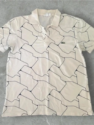 Vit mönstrad pikétröja från Lacoste - Vit pikétröja från Lacoste med svart abstrakt mönster över hela tröjan. Klassisk krage, knappar vid halsen och den ikoniska gröna krokodilloggan på bröstet. Perfekt för en clean och sportig look. Snabb affär!