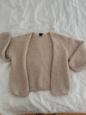 Beige stickad kofta från Lindex - Mysig beige stickad kofta från Lindex med öppen front och avslappnad passform. Koftan har kortare ärmar och är tillverkad i ett mjukt, fluffigt material som känns skönt mot huden. Perfekt att slänga på sig när det är lite kyligt ute.