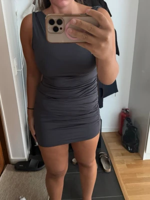 Mörkgrå tight kort klänning - Snygg mörkgrå kort klänning med tight passform och rynkade detaljer på sidan. Ärmlös modell med rund halsringning och slät baksida. Perfekt för dig som gillar en stilren och enkel look.
