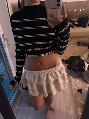 Svart cropped tröja med vita ränder - Säljer en svart ribbad cropped tröja med vita horisontella ränder. Tröjan har långa ärmar med tumhål och en hög hals. Materialet är stretchigt och mjukt, perfekt för en trendig och avslappnad look.